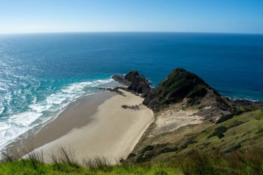 Kumlu bir sahil manzarası, kayalık çıkıntılar ve yemyeşil tepeler. Okyanus dalgaları, açık mavi gökyüzünün altında yavaşça kıyıya çarpar..