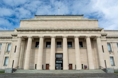 Yüksek sütunları ve geniş merdivenleri olan klasik mimari tarzı olan büyük bir bina. Gökyüzü parçalı bulutlu, derinlik katıyor..