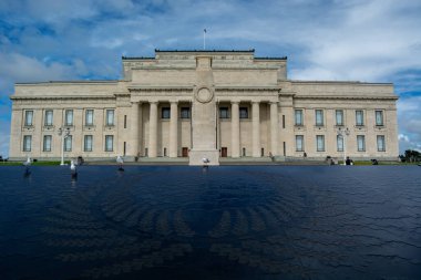Büyük merdivenleri ve sütunları olan büyük tarihi bir bina dağınık bulutlu mavi bir gökyüzüne karşı kurulmuş. Ön planda desenli bir yüzey ve yürüyen insanlar var..