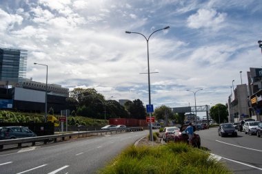 Araçların, sokak tabelalarının ve dağınık bulutlarla dolu temiz bir gökyüzünün olduğu kalabalık bir şehir yolu. Bölge yol kenarındaki binalar ve yeşilliklerle dolu..