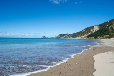 Yumuşak dalgaların sahile vurduğu, yemyeşil tepeler ve açık mavi bir gökyüzü olan sakin bir sahil sahnesi. Sahil şeridi sakin ve davetkar. Rahatlamak için ideal..