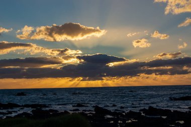 Okyanus üzerinde büyüleyici bir günbatımı, güneş ışınları bulutları aşıyor. Gökyüzü, suyun serin mavisiyle çelişen turuncu ve sarıya boyanmıştır. Ön plandaki kayalık kıyı şeridi sahneye doku ekler.