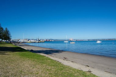 Huzurlu bir rıhtım sahnesi. Teknelerle dolu sakin bir körfez. Sahil şeridi açık mavi bir gökyüzünün altında çimen ve ağaçlarla kaplı. Uzakta endüstriyel yapılar ve dağlar görülebilir..