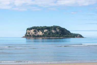 Berrak mavi gökyüzünün altında sakin sularla çevrili küçük bir adanın huzurlu manzarası. Ada, sükunet ve doğal güzelliği yansıtan yemyeşil ve kayalık uçurumlara sahip..