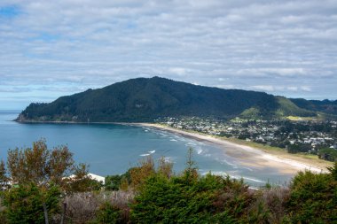 Kumlu bir sahil manzarası, dalgalı dalgalar ve yemyeşil bir yamaç. Gökyüzü parçalı bulutlu, sakin bir atmosfer yaratıyor. Kasaba kıyı şeridi boyunca görünür, ağaçların arasında yuva yapmış..