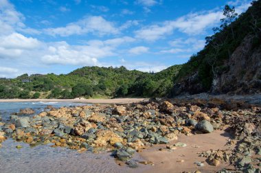 Kayalık arazi, yumuşak dalgalar ve parçalı bulutlu gökyüzünün altında yemyeşil tepeler içeren manzaralı bir sahil manzarası. Kıyı şeridi renkli taşlar ve çakıl taşlarıyla dolu..