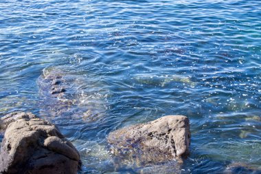 Kayalık oluşumlara hafifçe çarpan berrak mavi su manzarası. Güneş ışığı yüzeyde parlıyor, sakin ve davetkar bir atmosfer yaratıyor..