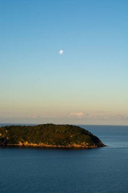 Berrak bir gökyüzünün altında sakin mavi sularla çevrili küçük, yemyeşil bir adanın olduğu sakin bir sahil manzarası. Ay gökyüzünde görünür, manzaraya sakin bir ortam ekler..