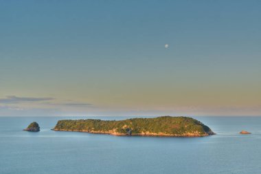Sakin bir adanın sakin bir görüntüsü, berrak bir gökyüzünün altında, görünür bir uyduyla çevrili. Manzara, adada yemyeşil ve uzak küçük adalarda bulunur..