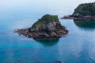 Sakin turkuaz sularla çevrili kayalık bir adanın bulunduğu sakin bir kıyı manzarası. Sahne, suyun yüzeyine yansıyan berrak gökyüzü ile doğanın sükunetini yakalar..