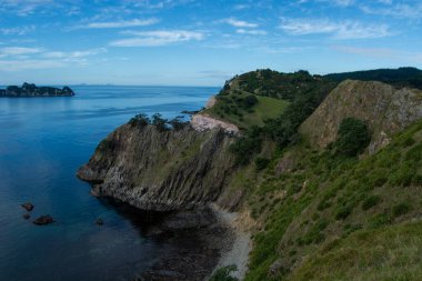 Berrak bir gökyüzü altında engebeli uçurumlar, yemyeşil yemyeşil suların ve sakin mavi suların bulunduğu manzaralı bir kıyı manzarası. Kıyı şeridi ağaçlar ve kayalık oluşumlarla kaplı, sakin bir doğal ortam yaratıyor..