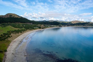 Sakin bir körfez ve kumlu sahilde hafif dalgaların dalgalandığı sakin bir kıyı manzarası. Arkaplanda yemyeşil tepeler yükseliyor parçalı bulutlu bir gökyüzünün altında barışçıl ve davetkar bir atmosfer yaratıyor..