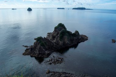Parçalı bulutlu bir gökyüzünün altında sakin mavi sularla çevrili kayalık adaları barındıran sakin bir kıyı manzarası. Sahne huzuru ve doğal güzelliği çağrıştırıyor..