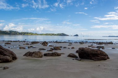 Pürüzsüz kum, dağınık kayalar ve sakin suların olduğu sakin bir sahil sahnesi. Arka planda, yeşil tepeler ve uzak bir ada mavi gökyüzünün altında tüylü bulutlarla görülebilir..