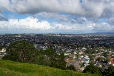 Tepenin tepesinden panoramik bir manzara, parçalı bulutlu bir gökyüzü altında yerleşim binaları ve yeşilliklerin karışımı ile genişleyen bir şehir alanını gösteriyor..