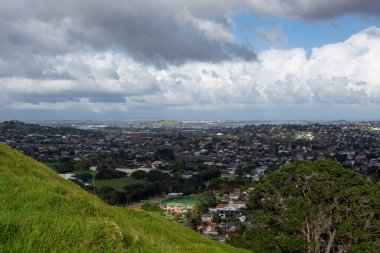 Yerleşim alanları, yeşil alanlar ve parçalı bulutlu bir gökyüzünün altındaki uzak tepelerin karışımından oluşan panoramik bir manzara. Ön planda yemyeşil çimenler ve büyük bir ağaç yer alırken, arka planda geniş bir şehir manzarası ve h yer almaktadır.