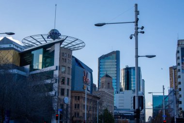 Modern gökdelenlerin ve üzerinde dairesel bir tasarım olan eşsiz bir mimari yapının yer aldığı bir şehir manzarası. Sahne, açık mavi bir gökyüzü altında çağdaş binalar ve şehir altyapısının bir karışımını yakalıyor..