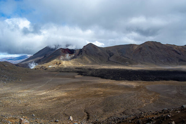 A vast volcanic landscape featuring a prominent volcano with a cloud-covered summit. The foreground shows barren land with rocky textures, while the background reveals rugged mountains under a cloudy sky.