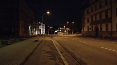 A deserted street at night with dim streetlights illuminating the road. Buildings line the street, casting shadows. The scene conveys a quiet, eerie atmosphere, typical of late-night urban settings.