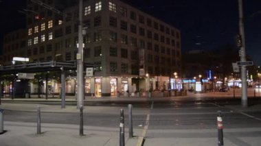 A quiet urban street at night, featuring modern buildings and illuminated storefronts. The scene captures a tranquil atmosphere with tram tracks visible and streetlights casting a soft glow.