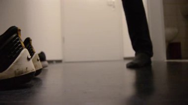 A person tying shoelaces on a pair of athletic shoes in a dimly lit room. The focus is on the shoes and hands, with a blurred background showing another pair of shoes nearby.