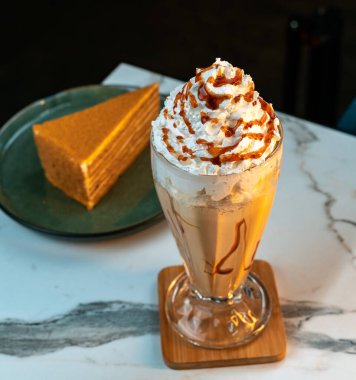 Çikolatalı milkshake, krem şantili ve uzun camlı karamel yağmurlu. Arka planda bulanık katlı kek dilimi. Tatlı menüleri, kafeler, yemek blogları ve yaşam tarzı içeriği için mükemmel..
