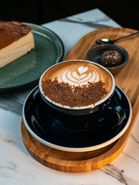 Çikolata parçacıklı kahve ve mantar ve ahşap tepside Bask peynirli kekle servis edilen kalp latte sanatı. Restoranlar, pastaneler, menü tasarımları, bloglar ve yiyeceklerle ilgili markalar için mükemmel..