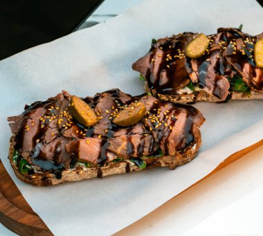 Pastırmalı gurme bruschetta, krem peynir, roka, kapari ve çoklu tahıllı tost üzerine balzamik jöle. Tahta tepside parşömen üzerinde servis edilmiş. Restoran menüleri, esnaf kafeleri, yemek promosyonları ve yemek tasarım projeleri için mükemmel..