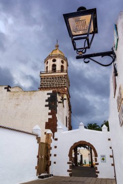 Teguise, Lanzarote, Kanarya Adaları, İspanya. Kilise kulesi manzarası, beyaz taş kapı ve bulutlu gökyüzüne karşı bir sokak lambası. Lanzarote 'nin tipik mimarisi.