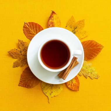 Sarı zemin üzerinde iki tarçın çubuğu ve renkli sonbahar yapraklı beyaz bir fincan siyah çay. Üst Manzara. Sonbahar sakin bir hayat.