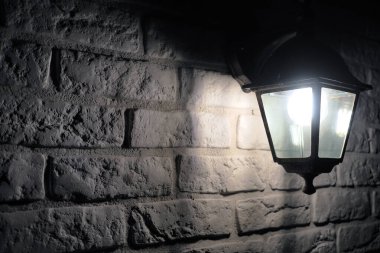 Retro-style lamp hangs on a white brick wall, copy space. Black metal wall lamp at night in the dark.