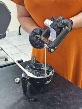 Siyah eldiven giyen bir kadın siyah bir kasede saç boyasını karıştırıyor. Profesyonel bir kuaför salonunda güzellik becerilerini gösteriyor. Özenli uygulama ve tekniğe odaklanmış durumda..