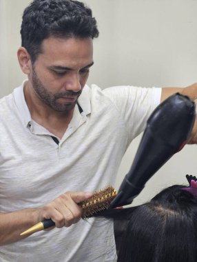 Bir kuaför müşterisinin saçını şekillendirmeye odaklanır. Güçlü bir saç kurutma makinesi ve yuvarlak bir fırça kullanarak onun kilitlerini şık ve sofistike bir görünüme dönüştürüyor. Ortam, bakım ve dönüşüm hissi veren bir güzellik salonu gibidir..