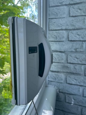 Arka planda yeşil ağaçlar olan cam bir yüzeye monte edilmiş bir robot pencere temizleyici cihazın yakın çekimi. Zahmetsiz pencere temizliği için akıllı ev teknolojisi. Yüksek kalite fotoğraf