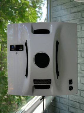 Konut ortamında cam bir pencereye bağlı otomatik cam temizleme robotu. Akıllı cihaz iş başında, güvenli bir şekilde bir iple korunuyor, dikey cam yüzeyini yeşil bir manzarayla temizliyor.