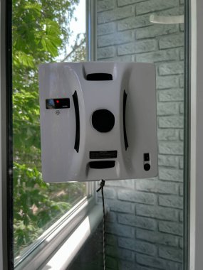 Konut ortamında cam bir pencereye bağlı otomatik cam temizleme robotu. Akıllı cihaz iş başında, güvenli bir şekilde bir iple korunuyor, dikey cam yüzeyini yeşil bir manzarayla temizliyor.