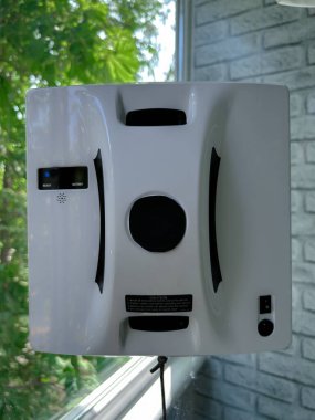Konut ortamında cam bir pencereye bağlı otomatik cam temizleme robotu. Akıllı cihaz iş başında, güvenli bir şekilde bir iple korunuyor, dikey cam yüzeyini yeşil bir manzarayla temizliyor.