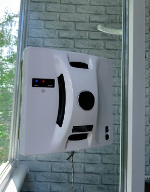Konut ortamında cam bir pencereye bağlı otomatik cam temizleme robotu. Akıllı cihaz iş başında, güvenli bir şekilde bir iple korunuyor, dikey cam yüzeyini yeşil bir manzarayla temizliyor.