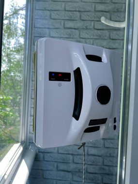 Konut ortamında cam bir pencereye bağlı otomatik cam temizleme robotu. Akıllı cihaz iş başında, güvenli bir şekilde bir iple korunuyor, dikey cam yüzeyini yeşil bir manzarayla temizliyor.
