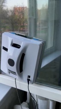 Konut ortamında cam bir pencereye bağlı otomatik cam temizleme robotu. Akıllı cihaz iş başında, güvenli bir şekilde bir iple korunuyor, dikey cam yüzeyini yeşil bir manzarayla temizliyor.