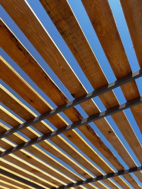 Açık mavi gökyüzüne karşı eşit aralıklı çıtalarla modern ahşap pergola çatısı. Mimari yapı, ışık ve gölgeli dinamik bir geometrik kalıp yaratarak doğal ahşap dokusunu vurguluyor.