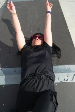 Sporcu kadın kolları havada, güneş gözlüğü takmış ve güneşin tadını çıkararak kaykay rampasında uzanıyor.