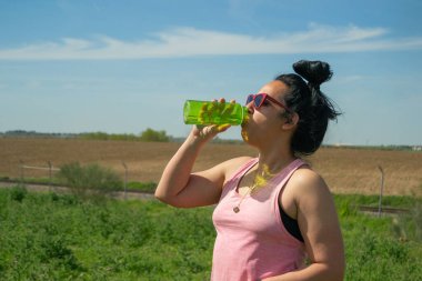 Baharda dışarıda egzersiz yaptıktan sonra yeşil bir şişeden su içen genç bir kadın.