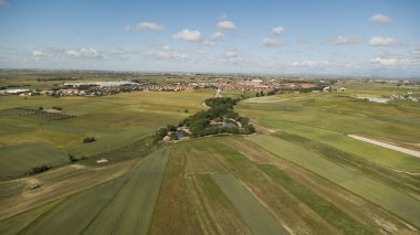 Kırsal bir arazinin havadan görünüşü ekilmiş tarlaları, küçük gölleri ve mavi gökyüzünün altında bulutlu bir köyü gösteriyor.