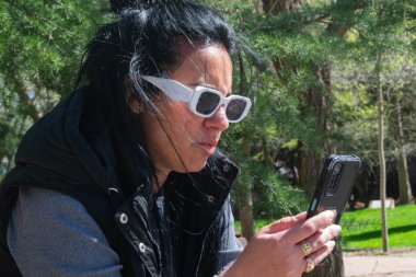 Kadın güneş gözlüğü takıyor ve parkta telefonunu kullanıyor.