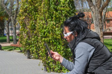 Güneşli bir günde yeşil bir çitin yanındaki bankta oturan bir kadın cep telefonunu kullanıyor.