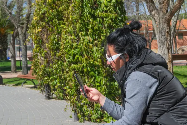 Güneşli bir günde yeşil bir çitin yanındaki bankta oturan bir kadın cep telefonunu kullanıyor.