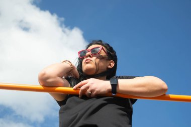 Sporcu bir kadın antrenmanına ara veriyor, yatay bir çubuğa yaslanıyor ve arka planda bulutlu mavi bir gökyüzü ile başka yere bakıyor.