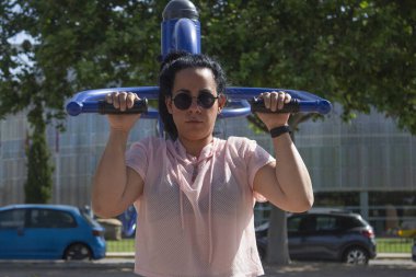 Parktaki mavi bir makinede destekli barfiks çeken kadın dayanıklılık ve kendini spora adamış.