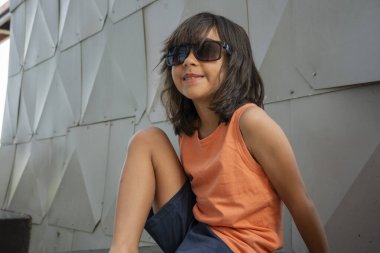 Yazın modern metal bir duvarın yanında oturan, güneş gözlüğü takan gülümseyen genç bir çocuğun portresi.